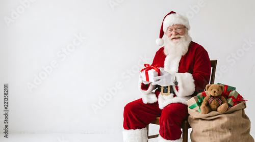 Smiling Santa Claus in traditional red suit sitting on a chair next to a large sack filled with wrapped gift boxes and a teddy bear, holding a small present against a white background.