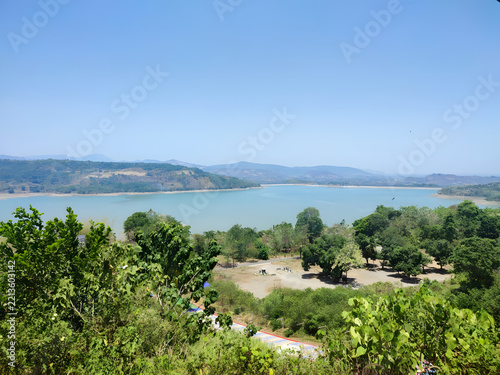 High-angle view of a calm lake framed by vibrant green vegetation, rolling hills, and a clear blue sky on a bright sunny day.