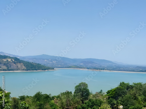 A tranquil turquoise reservoir stretches toward distant, hazy mountains under a clear blue sky, framed by lush green trees in the foreground, evoking a sense of peaceful natural beauty.
