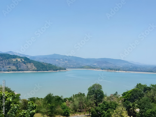 A wide shot capturing a calm lake surrounded by lush green trees and distant blue mountains under a clear sky.