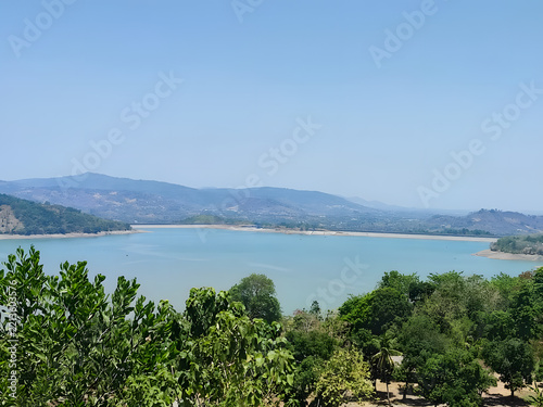 A tranquil reservoir with turquoise water stretches toward distant, hazy mountains under a clear blue sky, framed by lush green trees in the foreground, evoking a sense of calm and natural beauty.