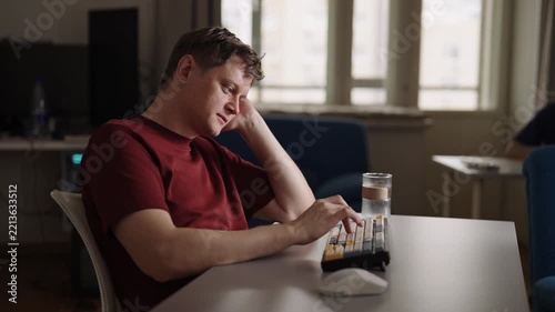 Tired young adult man feeling bored and uninspired at home office desk, props head on hand, procrastinating and struggling with creative block, burnout. Apathetic freelance worker, slow motion.