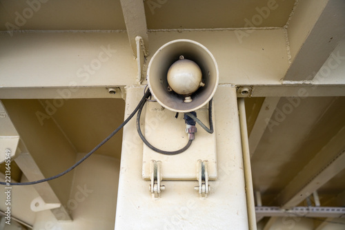 A horn loudspeaker with exposed cables is mounted on a pale metal support, viewed from below.