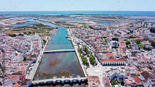Drone footage showcasing the historic roman bridge and the gilao river dividing the charming coastal town of tavira, revealing the scenic landscape and salt pans leading to the atlantic ocean
