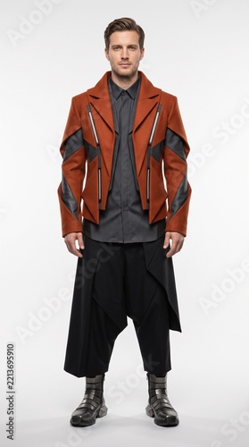 Man wearing a futuristic terracotta rust jacket, dark shirt, and black wide-leg trousers isolated on white.