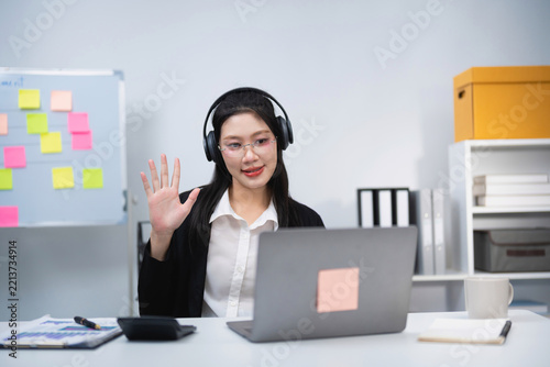 Young woman wearing headphones waving hand during video call on laptop in modern office with whiteboard and shelves