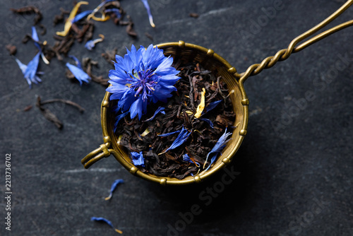 Loose Earl Grey tea leaves with cornflower blossom in brass tea infuser. Dark marble background. Close up. Top view.