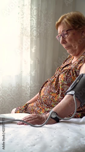 An elderly woman sitting on the bed, taking her blood pressure with an electronic blood pressure monitor