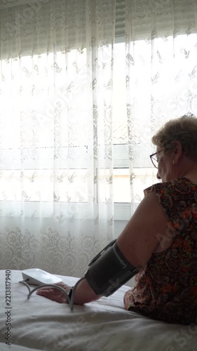 An elderly woman sitting on the bed, taking her blood pressure with an electronic blood pressure monitor
