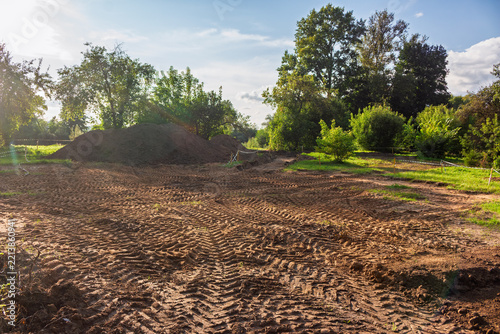 Heavy construction machinery tracks on soil during park renovation work, showing a large dirt mound and cleared land with trees in the background on a sunny day