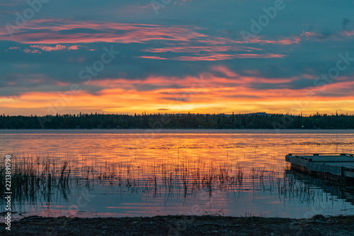 Orange sunset reflected in calm lake water with reeds and fishing pier