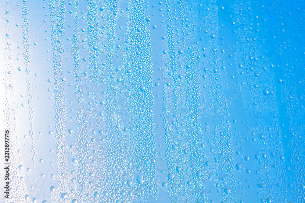 Fototapeta premium Water droplets bead across a glass surface, creating vertical trails and sparkling textures. Bright light gives the cool, abstract image a fresh, translucent appearance