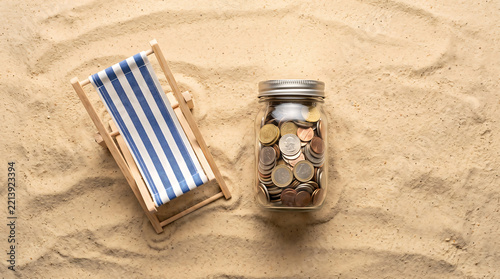 A miniature wooden beach chair with blue and white stripes placed next to a glass jar filled with assorted metal coins on a textured sandy surface