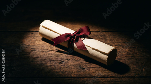 A rolled parchment scroll tied with a dark red ribbon, resting on a dark, rustic wooden surface illuminated by a single spotlight from above