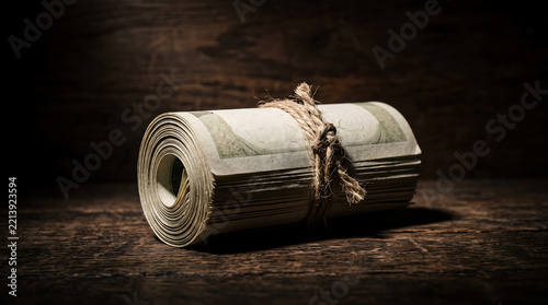 A rolled-up stack of united states dollar bills, tied with a piece of brown twine, resting on a dark, rustic wooden surface with dramatic side lighting