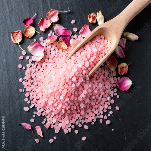Coarse pink Epsom bath salts in wooden scoop with rose petals, holistic wellness therapy promotion background.