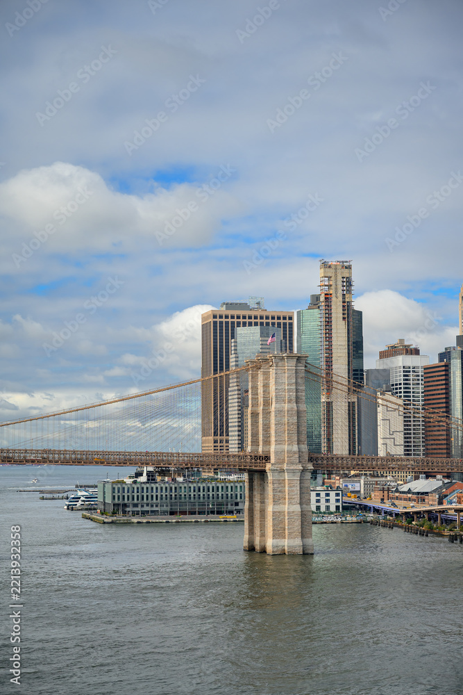 Fototapeta premium A tower of Brooklyn Bridge rises beside the Manhattan waterfront. Stone arches overlook gray river water with a glass pier building below a clouded blue sky.