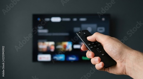 Hand holding remote control pointing at smart TV screen with streaming apps.
