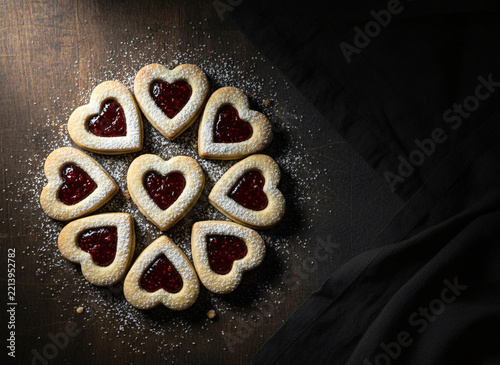 Heart-shaped linzer cookies with red jam, arranged with festive care, moody dark background with a single warm highlight