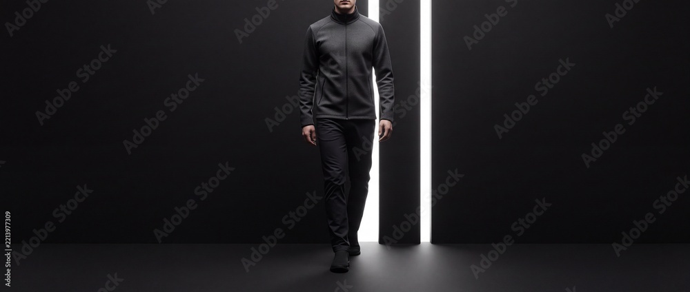 Naklejka premium Confident man in sleek athletic techwear walking forward through a dark minimalist space with vertical light bars, representing a metaphor for progress, modern urban fashion, and editorial style.