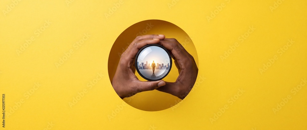 Fototapeta premium Hands holding a round lens showing a businessman walking towards a modern city skyline at sunset, seen through a hole in a yellow background, symbolizing business vision, future goals, and career