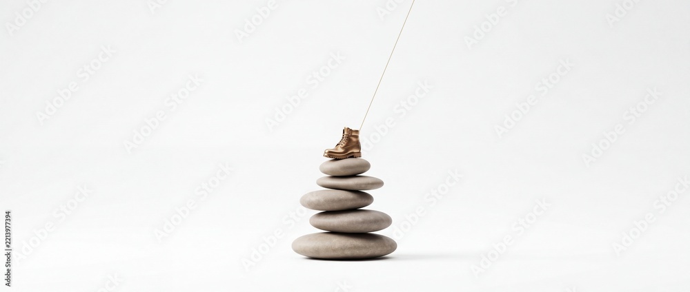 Fototapeta premium Conceptual image of a golden miniature work boot perfectly balanced on a stack of smooth grey zen stones with a thin wire against a white background, symbolizing business stability and career growth.
