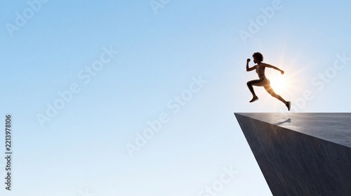 Silhouette of a motivated young woman jumping off a concrete ledge into the clear blue sky with large negative space on the left side, representing freedom, courage, and taking a leap of faith.