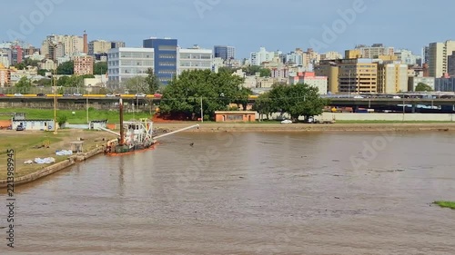 Water dredging vessel operating near a lush riverbank in Porto Alegre Brazil with a wide urban backdrop of highways, overpasses, and city buildings under a blue sky