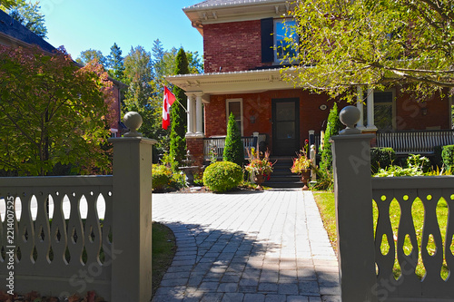 Walkway through a decorative gate to a red brick house with a Canadian flag on the porch, on a sunny autumn day