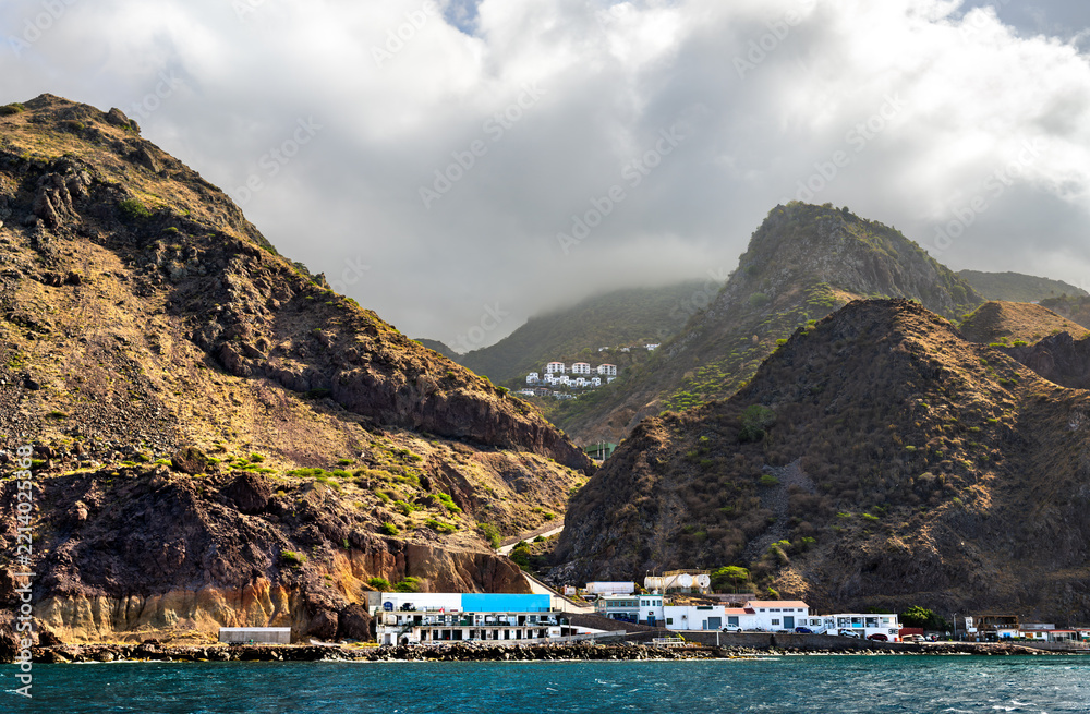 Fototapeta premium Scenic view of the rocky coast, Fort Bay harbor buildings, and steep volcanic mountain slopes on the tropical island of Saba in the Dutch Caribbean under a cloudy sky.