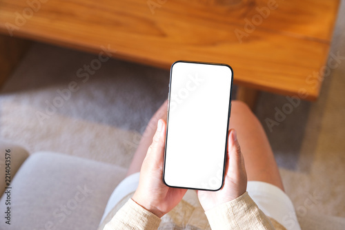 Top view mockup image of a woman holding and using mobile phone with blank desktop white screen at home