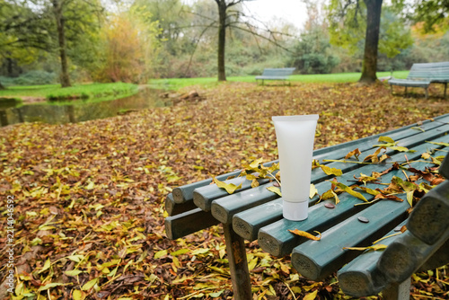 Cosmetic product tube standing on park bench outdoors in autumn surrounded by fallen leaves, trees and tranquil seasonal landscape