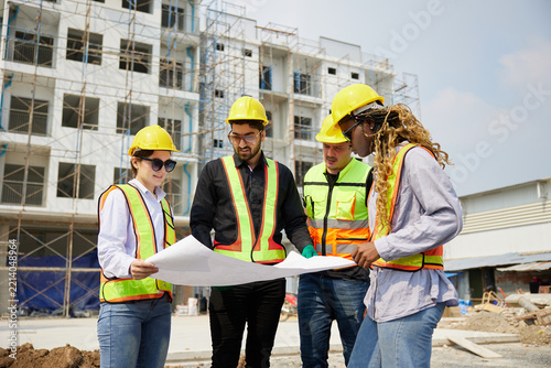 Group of construction workers, wearing hard hats and safety vests, are gathered around a blueprint at a construction site.