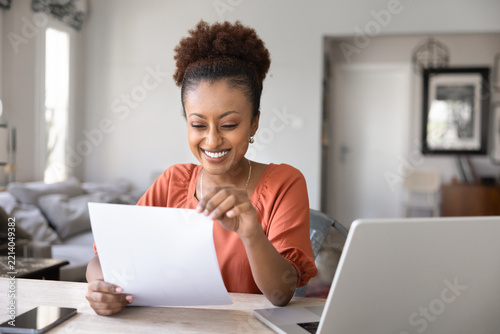 Cheerful African woman reading printed document seated at desk with laptop and smartphone, smiling, enjoying professional or personal achievement, bank loan acceptance, reward or great business news