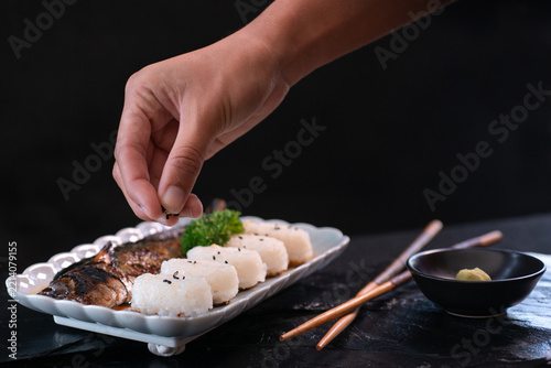 A chef's hands are arranging a ready-to-eat Japanese meal consisting of rice balls and grilled mackerel, accompanied by chopsticks and a small cup of wasabi arranged side-by-side on a black stone surf