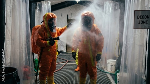 Emergency personnel in orange hazmat suits undergoing decontamination with water spray in a decon zone