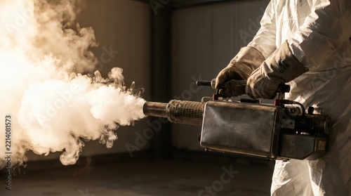 Professional worker in protective suit operating a fogging machine, emitting thick smoke for disinfection, pest control, or sanitization in an indoor environment.