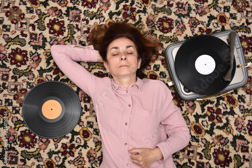 Nostalgic senior woman with closed eyes lying on a Persian carpet enjoying listening to music on a vintage record player. Top view. Concept of people, vintage objects and nostalgic moments.