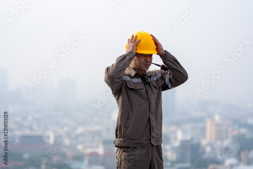 Construction worker wearing yellow helmet in protective work clothing against hazy urban skyline, showing focus and readiness at elevated outdoor worksite