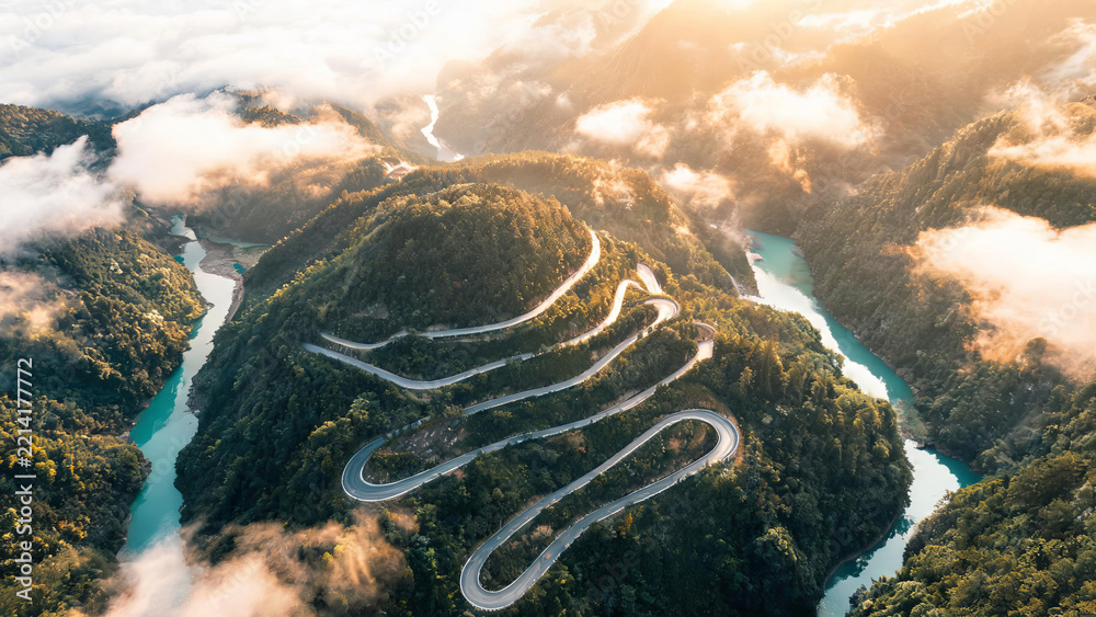 Naklejka premium An aerial view of a winding mountain road cutting through forested hills beside turquoise rivers, with clouds drifting in valleys at sunrise.