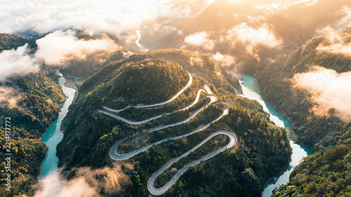 An aerial view of a winding mountain road cutting through forested hills beside turquoise rivers, with clouds drifting in valleys at sunrise.