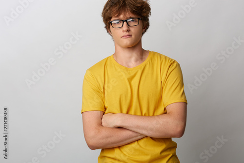 Obraz na plátně Young man with curly hair in glasses wearing yellow t-shirt standing with arms crossed on gray background