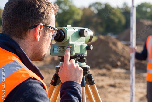 Land Surveyor in Orange Safety Vest Using Optical Level on Tripod at Construction Site