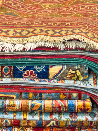 Colorful stacked traditional rugs with intricate patterns and fringes displayed in a textile shop, showcasing rich craftsmanship and design
