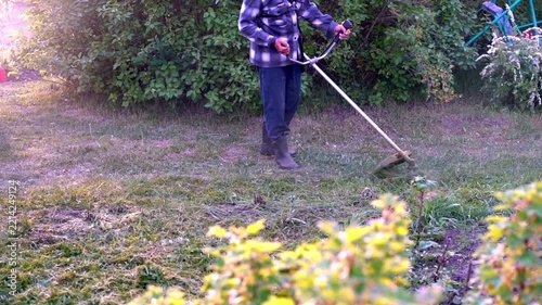 Sunlight glints off the grass as the person cuts it. They move steadily, wearing boots and a plaid shirt. Gardens and shrubs frame the scene behind them. They focus, tending to their yard with purpose