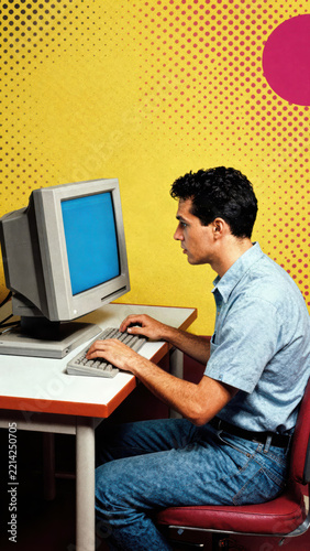 Young man curly hair blue shirt sitting typing vintage computer CRT monitor retro desk yellow background pop art focused expression