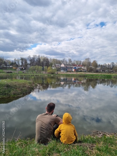 Obraz na plátně Vertical back view of a father and child sitting on a grassy river bank, looking