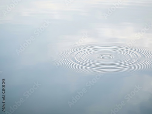 Concentric circular ripples expanding across a calm, smooth water surface, creating a peaceful and minimalist effect with soft light reflection.