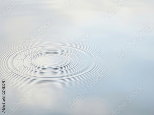 Concentric ripples spreading across a calm, smooth water surface, creating a peaceful and minimalist pattern with soft reflections of a pale sky.
