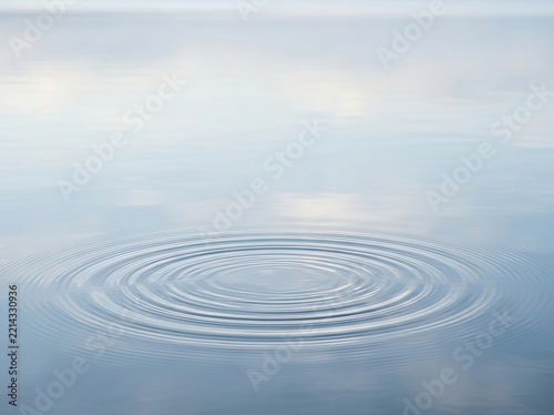 Concentric ripples spreading across the calm, reflective surface of clear blue water with a soft, blurred horizon.